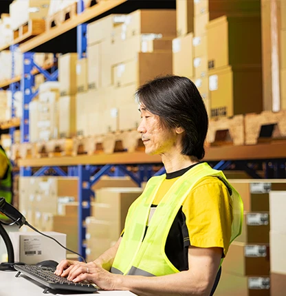 Warehouse worker booking stock in at a keyboard with a barcode scanner, cartons on the racking behind